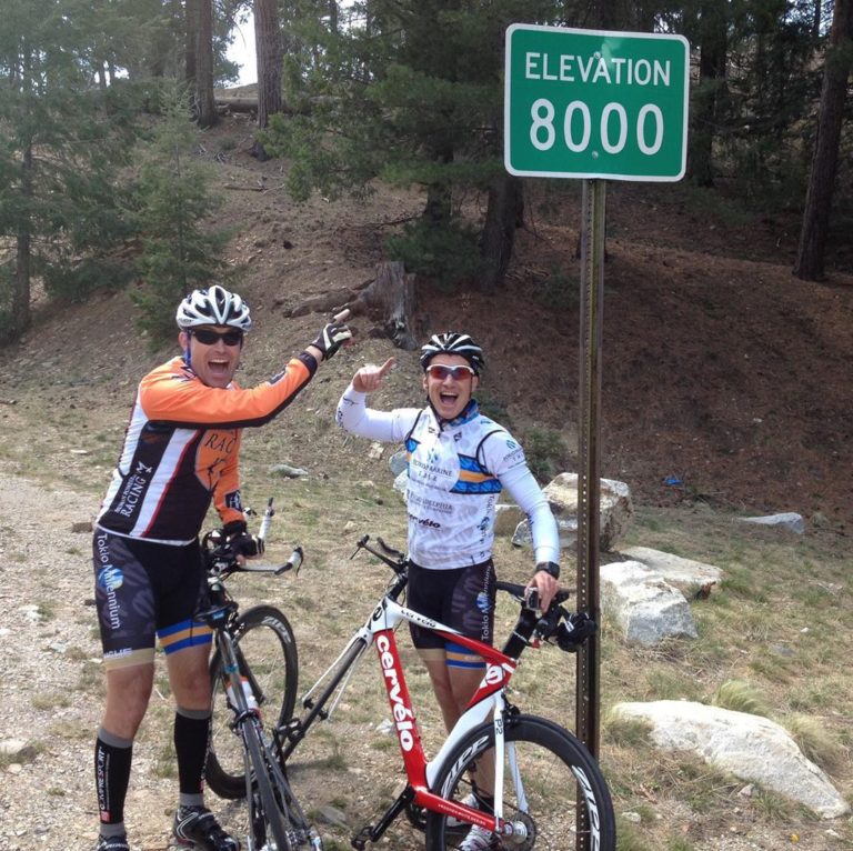 A green rectangular sign on a black pole Elevation 8000 stands on a trail at the edge of trees and beside white rocks on the ground. Two cyclists in helmets and gear with their bikes point to the sign with smiling faces.
