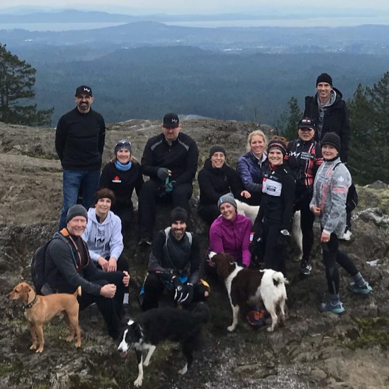 About a dozen people in winter gear pose with three dogs on a mountain top with blue haze of hills and sky in the background