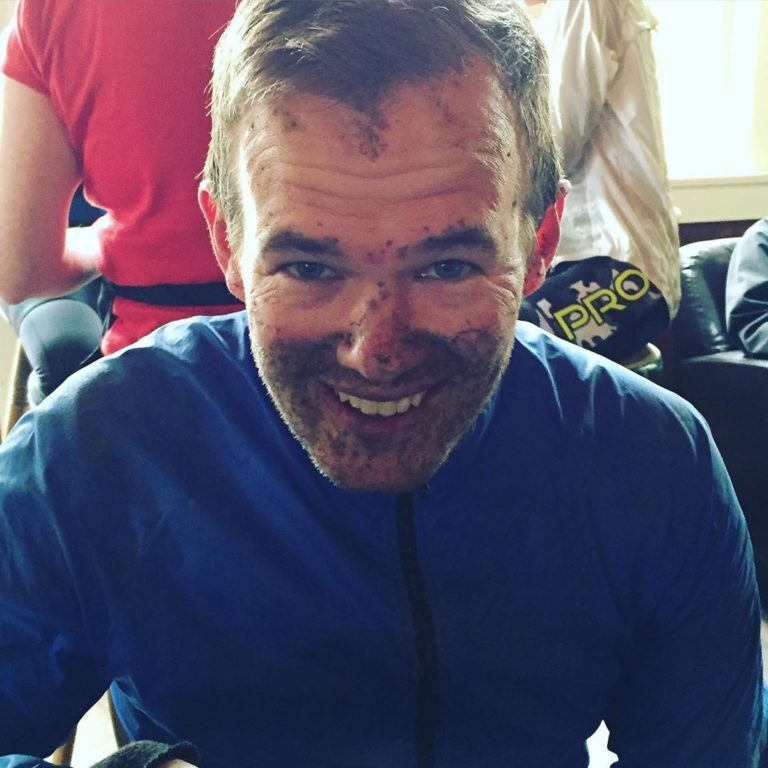 Close up of the face of a male adult smiling at the camera, in a blue biking t shirt