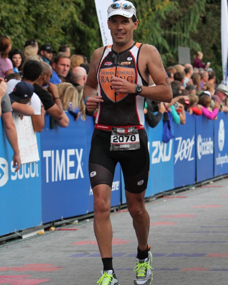 One male runner in black athletic gear is confidently in mid stride along a race on a road while people watch from the side behind a blue barrier