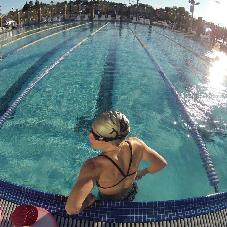 Female swimmer wearing a swim cap and swim suit has back and shoulders visible to camera, standing in an empty pool with the lanes stretching out in front her in blue water.