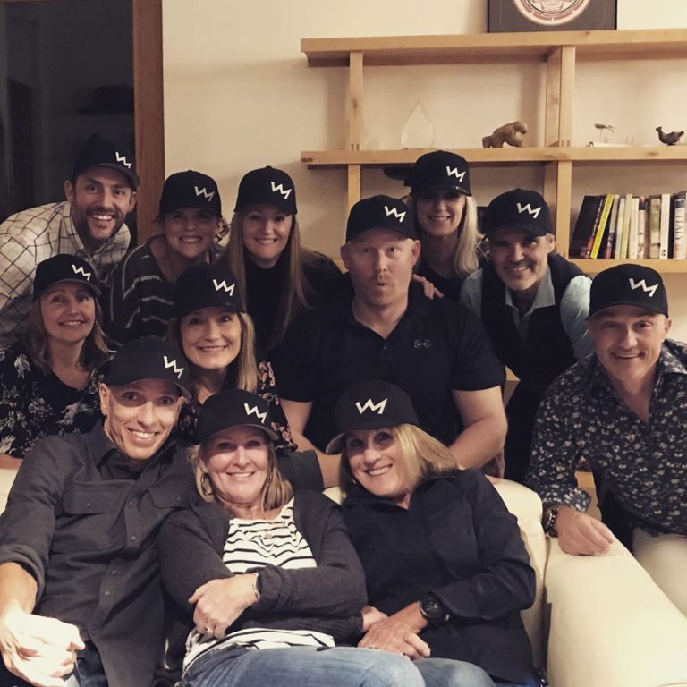 About a dozen men and women pose with black hats with a lightning bolt on it in a living room, with three in a big white chair and the others assembled in fron to fa bookcase behind the chair