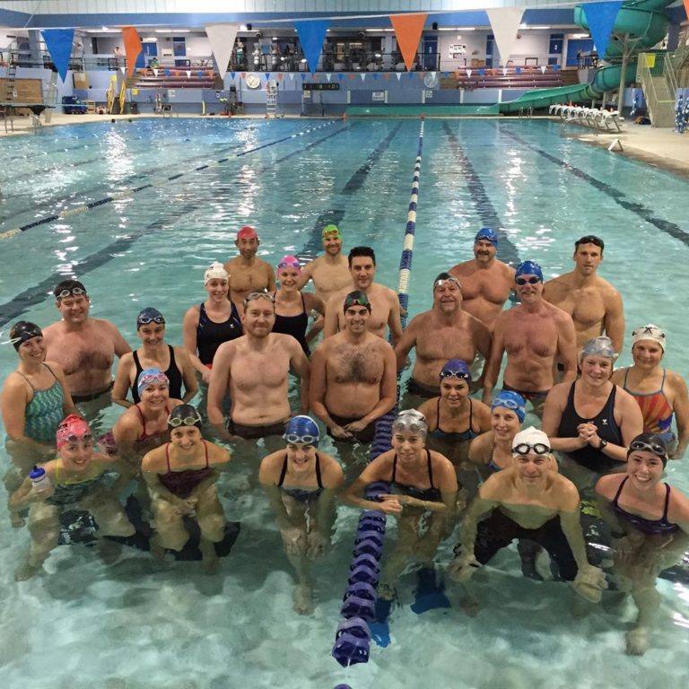 About a dozen people in swim wear pose together in a pale blue indoor pool