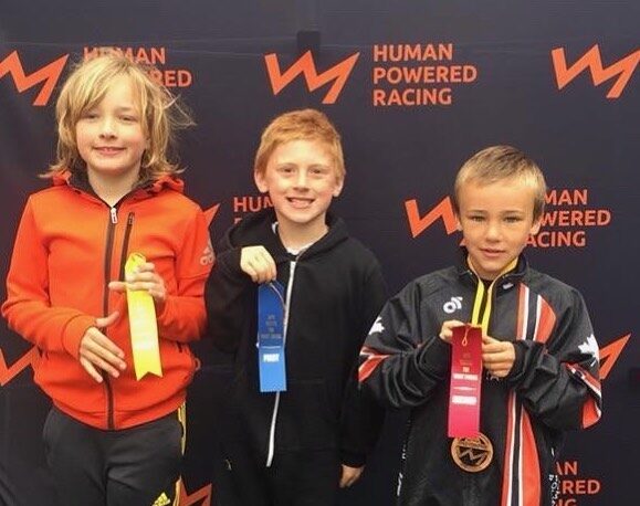 Three young boys stand in a row in front of a black banner with Human Powered Racing written on it and its lightning bolt logo. They wear or hold medals each in gold, blue and red.