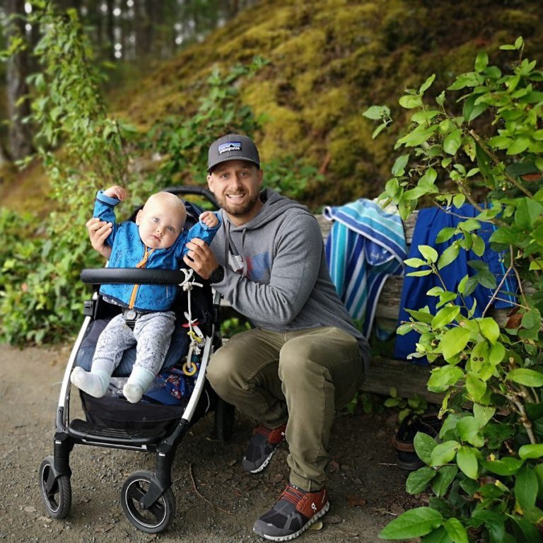 baby in a blue and grey outfit in a stroller and an adult, perhaps the father holds the baby's arms up to indicate cheering. Adult male is wearing a grey hat and grey sweatshirt with olive green pants. He is sitting on a bench with two blue towels hanging.