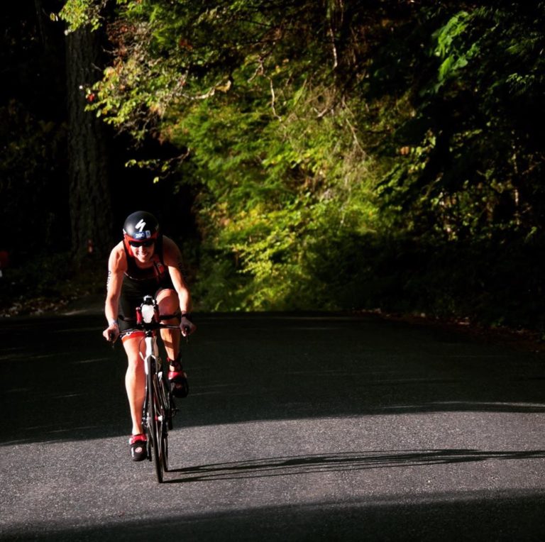 One athlete in biking gear on a bike on a grey wide road with the bike's long shadow visible across the road, passing under the low lying branches of a green tree