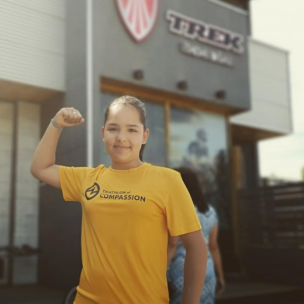 A child of about 13 years curls their arm up to show their muscles, wearing a yellow T shirt and smiling at the camera. She is standing in front of a store front that says Trek