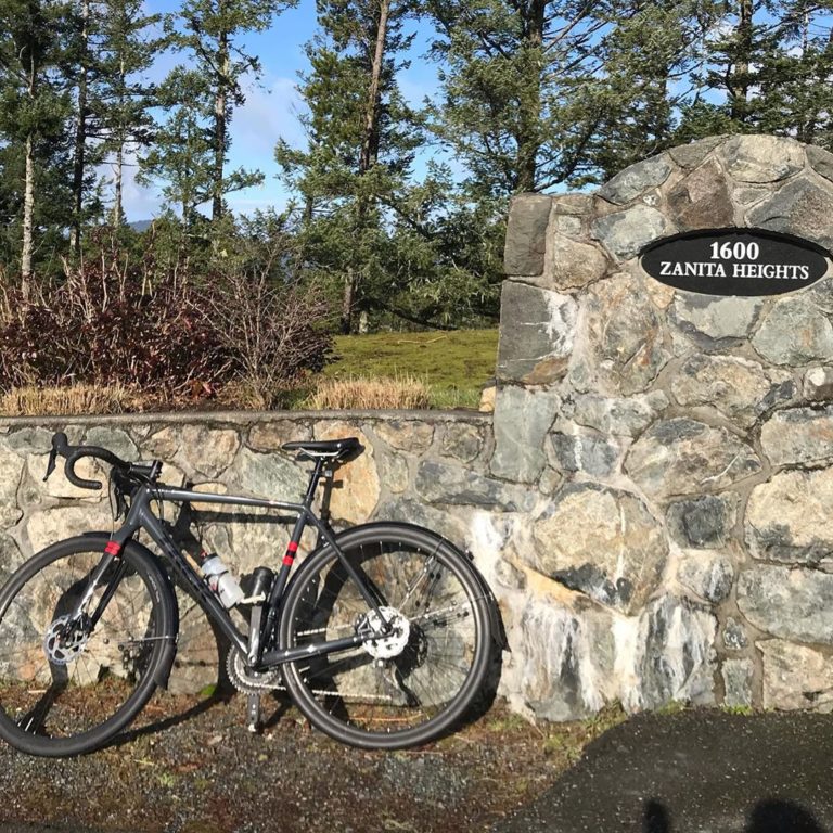 Bicycle parked beside a rock wall with a black oval sign Zanta Heights on it, some fir trees and blue sky visible behind