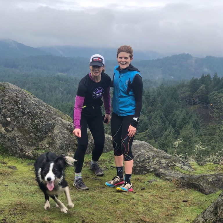 Black and white border collie stands with its human and their friend on a mountaintop. Two women in hats and blue and pink lined black running gear stand posing.