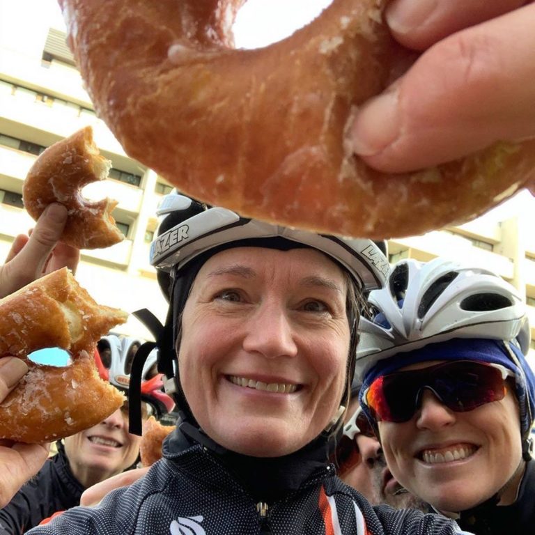 Two bikers visible in close up with helmets on and big grins on their faces, while a few more bikers and donuts partially eaten can be seen in the background. One donut is visible in extreme close up