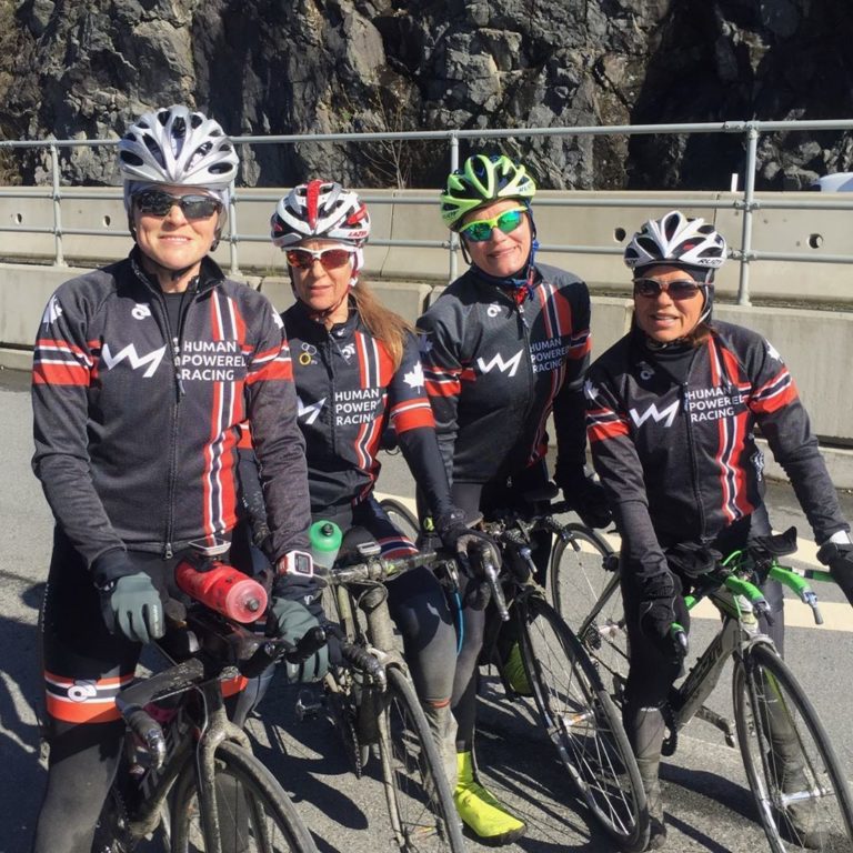 Four bikers on their bicycles and wearing running gear and helmets pose on a road with a mountain cliff background