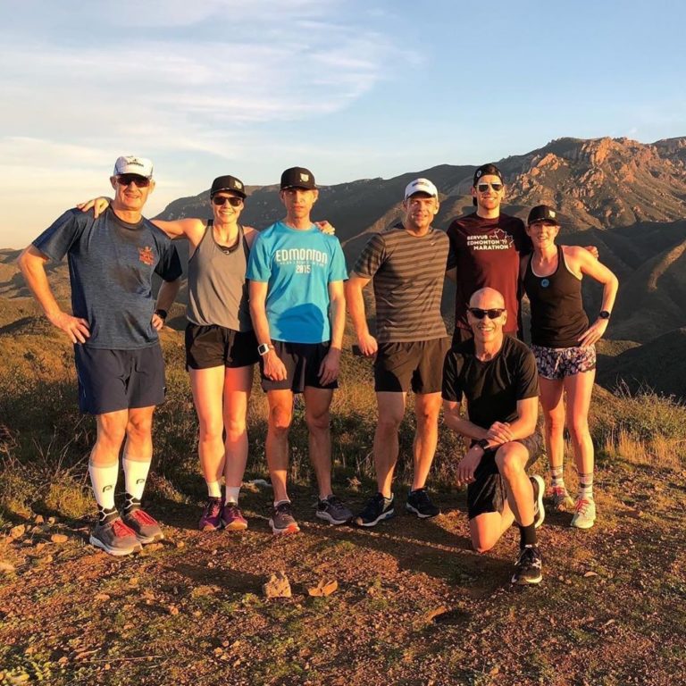 A half dozen male and female athletes in hats and summer running wear stand on a red brown summit with other mountains visible behind them under a bright blue sky.