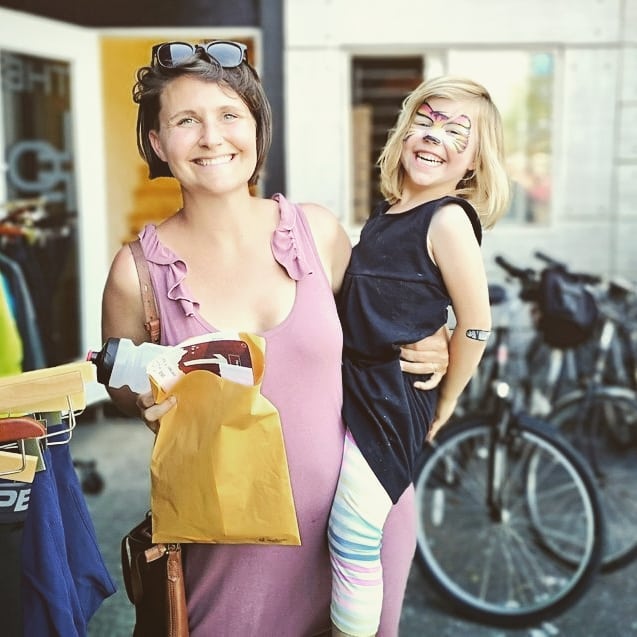 A woman in a pink dress with glasses perched on her dark brown hair holds up an open brown paper package and smiles at the camera. She is holding her daughter on her hop and standing in front of a bicycle store, with parked bikes and the store window visible blurrily in the background. The child is wearing a black top and pale multi coloured leggings. The child has face paint around her eyes and the elbow