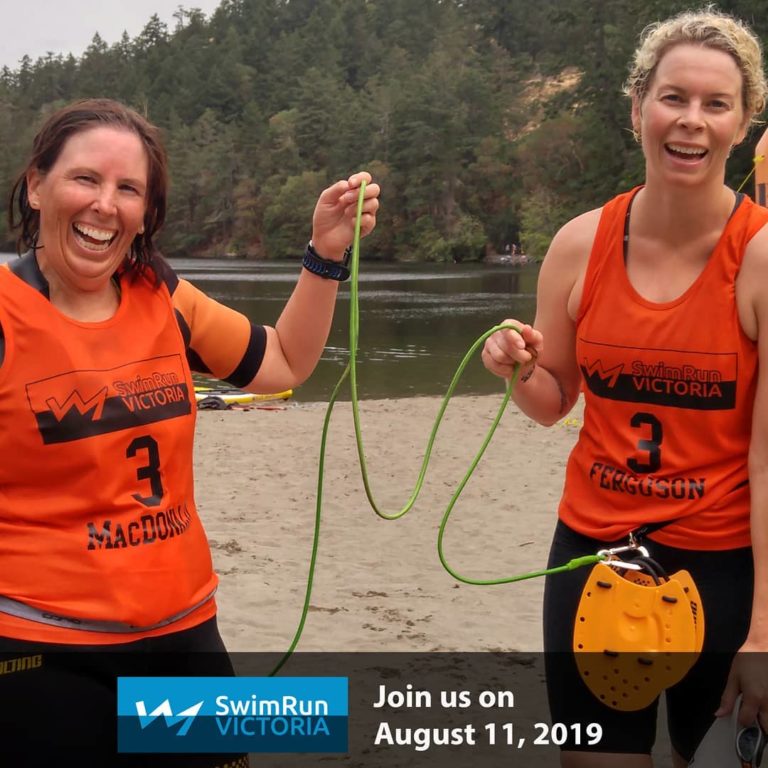 There is a banner in black at the bottom of the photograph that says Join us on August 11. 2019. Two women in red and black athletic gear with SwimRun Victoria written on it stand on a sandy beach by a lake with a hill of trees as background on the other shore. They hold a green line strung between their yellow paddles hanging on their waist and grin at the camera.