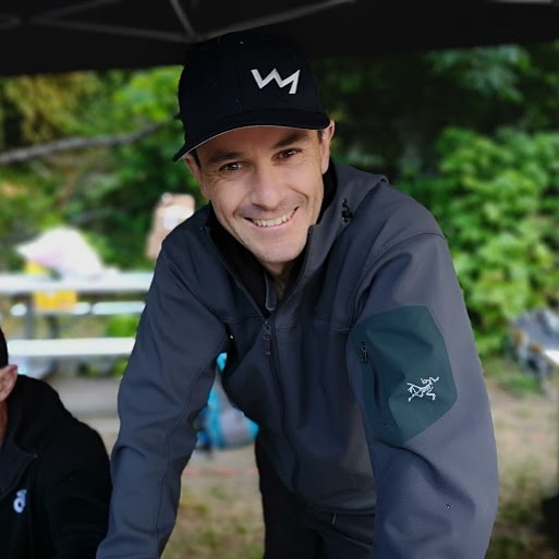 A smiling young man in a black hat with a lightning bolt logo and dark blue jacket poses on an exercise bike (off camera) outside, green trees in the back.