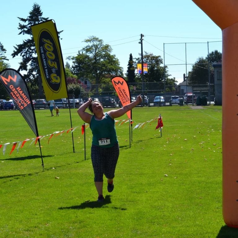 A runner with the tag 211 on their green t shirt is almost at the finish line. The orange arch is partially visible on the side of the photograph, ona sunny day on green grass.