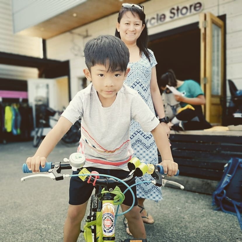 A child in a white T shirt sits astride a bicycle looking straight at the camera seriously while his mom is partially visible behind him smiles on. They are standing in front of a store.