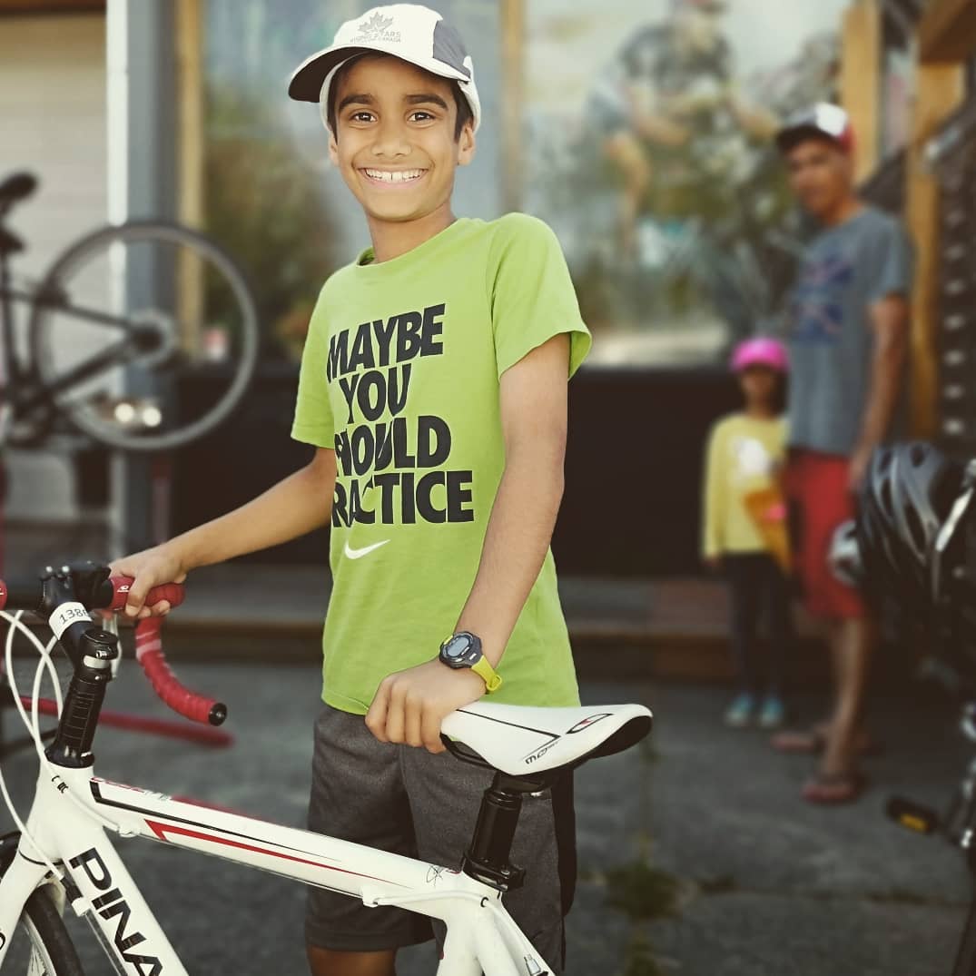 A child, perhaps 11 or 12 years of age stands beside a white bike that is partially visible. His parent and a younger sibling stand out of focus in front of a bike store window in the background. The kid has on a white hat and a green T shirt that has Maybe You Should Practice written on it.