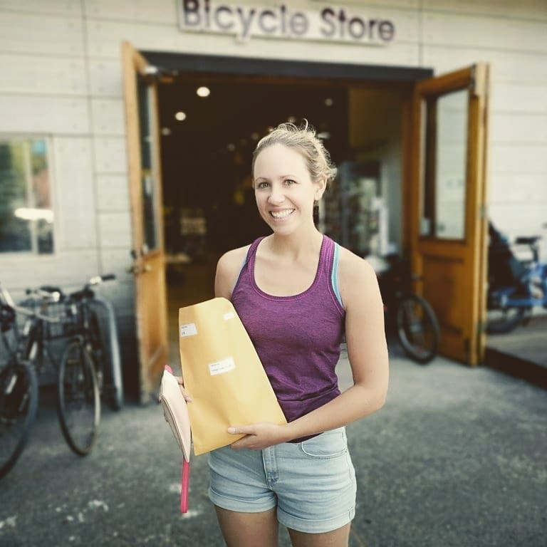 A woman in a dark purple tank top and light blue denim shorts stands in front of a building that says Bicycle Store, with some bikes parked in front of it. She holds up an open brown paper package and grins at the camera