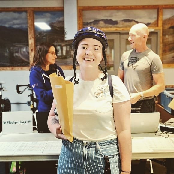 A young woman in a black bicycle helmet stands inside a building holding an open brown paper package. She is smiling, and standing in fron tof a table with a placard that says Pledge Drop Off. There are a man in beige and a woman in dar blue standing behind the table turned towards each other in middle of a conversation