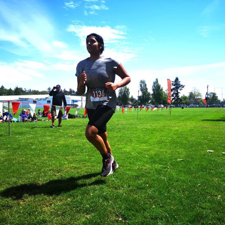 A young woman athlete runs the last part of the race on green grass, wearing grey top and black shorts and the number 1136 tagged on a white rectangle pinned to their clothes. The sky is blue and spectators watch
