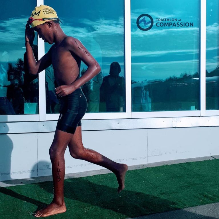 A young kid with a yellow swimmer's cap with the legend Human Powered Racing on it holds the cap with one hand on his head while running. He is wearing black swim shorts and behind him is a window reflecting the blue sky with the legend Triathlon of Compassion and a lightning logo inside a circle beside it