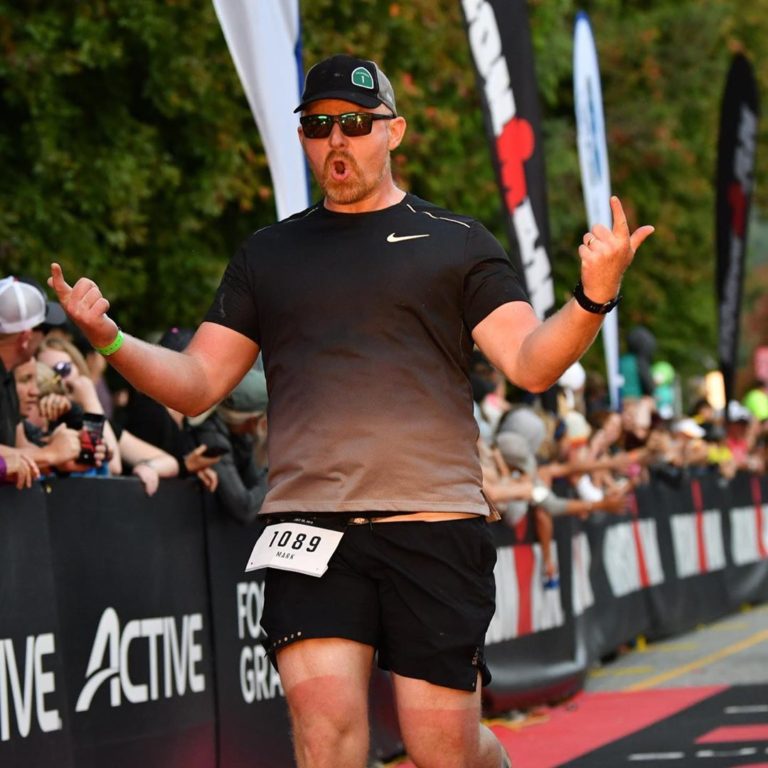 A male athlete completes a race, people lined up on the side of a black banner with advertising on it including the word 'Active' cheer him on. He wears a black hat, a darg grey t shirt and black running shorts with a rectangular patch 1029 on the shorts and has both hands raised to the side in jubilation