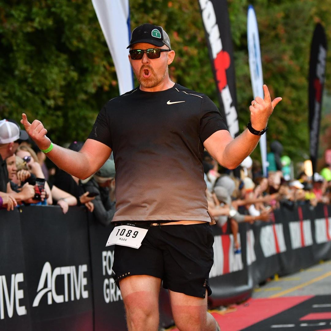 A male athlete completes a race, people lined up on the side of a black banner with advertising on it including the word 'Active' cheer him on. He wears a black hat, a darg grey t shirt and black running shorts with a rectangular patch 1029 on the shorts and has both hands raised to the side in jubilation