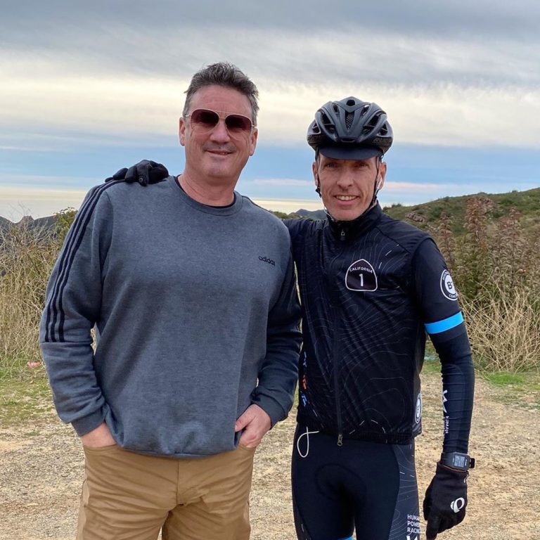 A man in black biking gear and a black helmet stands with his arm around the shoulder of another man in sunglasses, grey full sleeved T shirt and khakis under blue skies on a hill top
