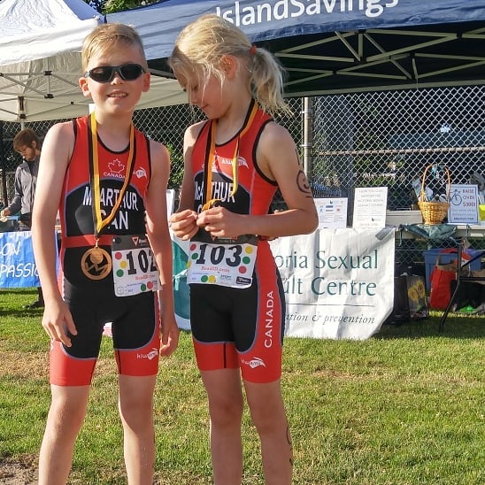 Two children stand side by side in red and black racing gear wearing rectangular race numbers on their waists. The boy is wearing dark sunglasses on his short blond hair and facing the camera with a smile. The girl is facing her her friend and has her longer blonde hair in a ponytail. A table with a white banner is visible blurred in the back ground behind the green grass.