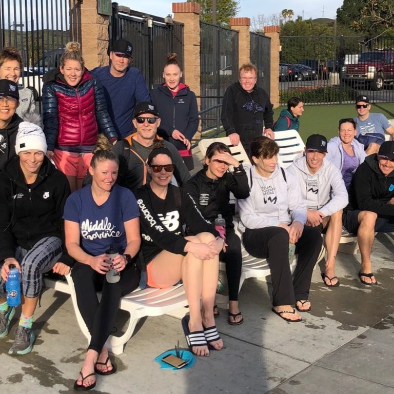 A dozen athletes in various exercise gear sit on two levels of steps by the side of a road beside a black grilled tall fence on a sunny day