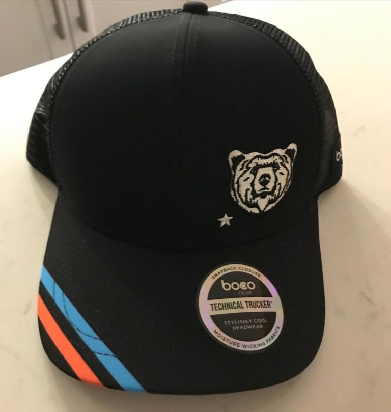 Black hat is displayed on a beige desk top. Hat has two parallel stripes in red and blue on the side of the visor, a circular logo with 'technical trucker' printed in the front of the visor and an image of a tiger head on the front of the dome.