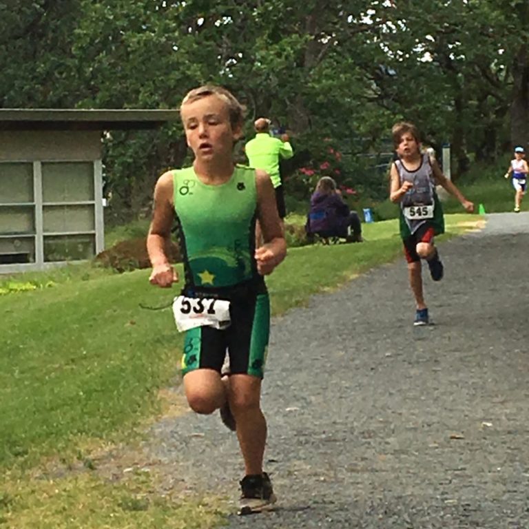 Young child of about 9 years wearing a green running top and black shorts with blue stripe is running, caught mid stride by camera with one knee raised. Another young kid is visible in the distance. The boys are running on a gravel trail with grass to the side and trees visible in the distance.