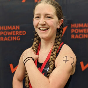 A youth racer flexes and makes a muscle in front of the Human Powered Racing championship board.