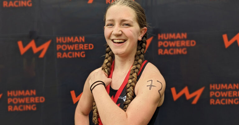 A youth racer flexes and makes a muscle in front of the Human Powered Racing championship board.