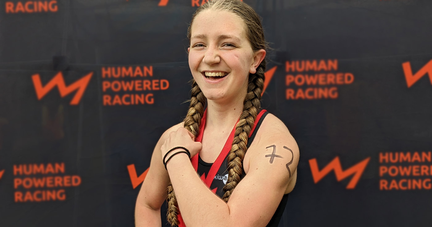 A youth racer flexes and makes a muscle in front of the Human Powered Racing championship board.