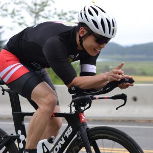 A Cowichan Challenge triathlete tucks on his bike along a straight stretch smiles broadly for the camera.