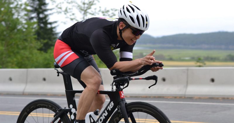 A Cowichan Challenge triathlete tucks on his bike along a straight stretch smiles broadly for the camera.