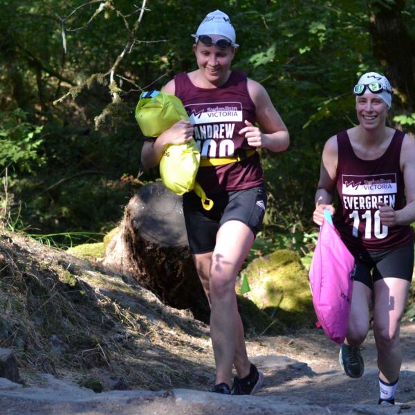 Two athletes race together, smiling, through the forest trail on their way to the next swim leg.