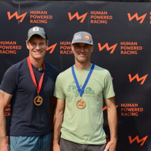 two men wearing medals standing in front of a Human Powered Racing media backdrop