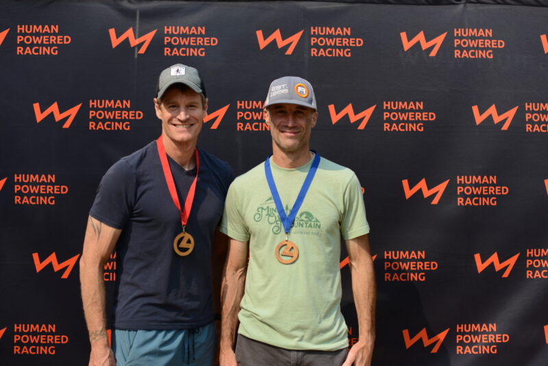 two men wearing medals standing in front of a Human Powered Racing media backdrop