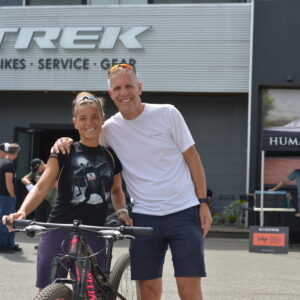 A racer and her partner pose below the TREK sign with her bicycle.