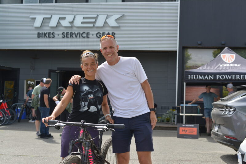 A racer and her partner pose below the TREK sign with her bicycle.